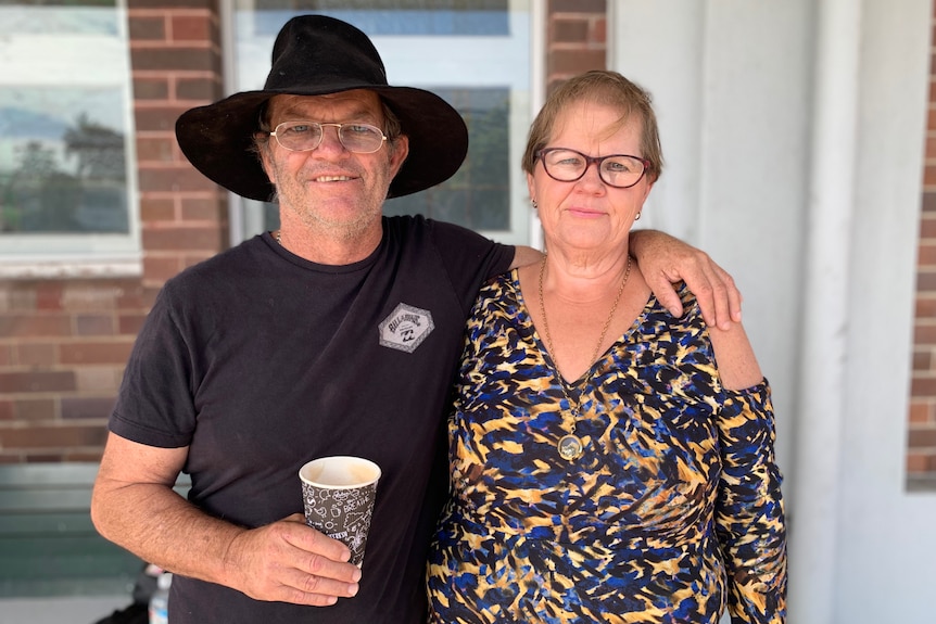 A man holding a cup of coffee with his arm draped over the shoulders of a woman looking unsmiling at the camera.