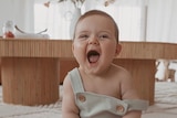 A baby sits on a rug wearing a onesy with a big smile. 