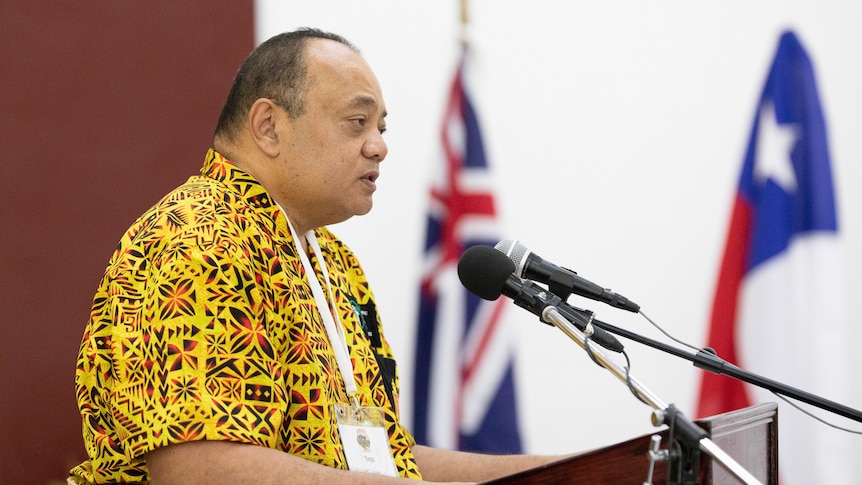 Tongan prime minister Siaosi Sovaleni, wearing a yellow, black and orange island shirt speaks at a microphone. 
