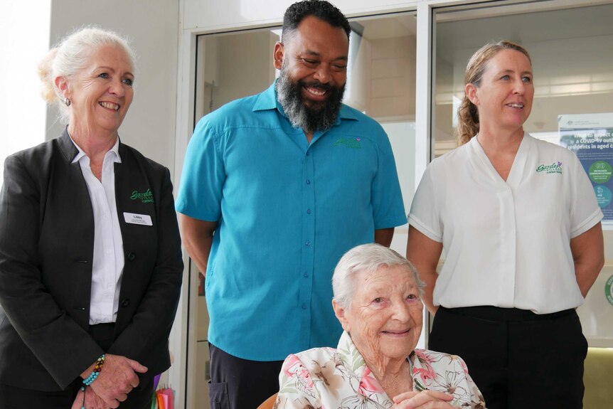 Three Garden Village staff stand behind 100 year old Grace who sits in front, all smiling.