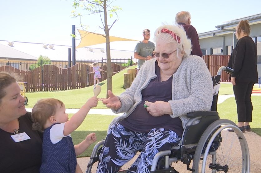 A elderly lady in a wheel chair interacts with a toddler, who is holding a maraca, while an childcare worker watches