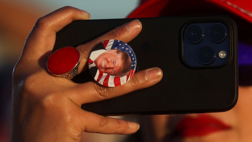 A close up of a person holding a smartphone with a Donald Trump button on the back 