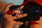 A close up of a person holding a smartphone with a Donald Trump button on the back 