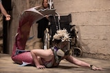A woman in her mid 30s in a mermaid costume and oxygen mask crawls away from a wheelchair