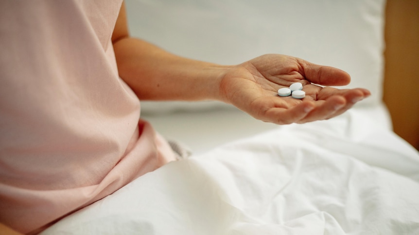 A woman in a peach coloured hospital gown holds three white pills in her left hand