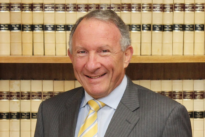 Gavin Silbert, in grey pinstriped suit, yellow tie and white and blue striped shirt, standing in front of a shelf law books