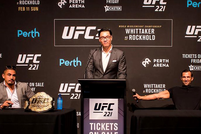 A commentator is flanked by two men at a media conference with UFC signage