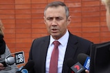 Roger Cook speaks into reporters' microphones in front of an orange-brick wall.