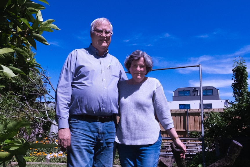 An older couple stand in their backyard with blue sky above and new apartments behind them.