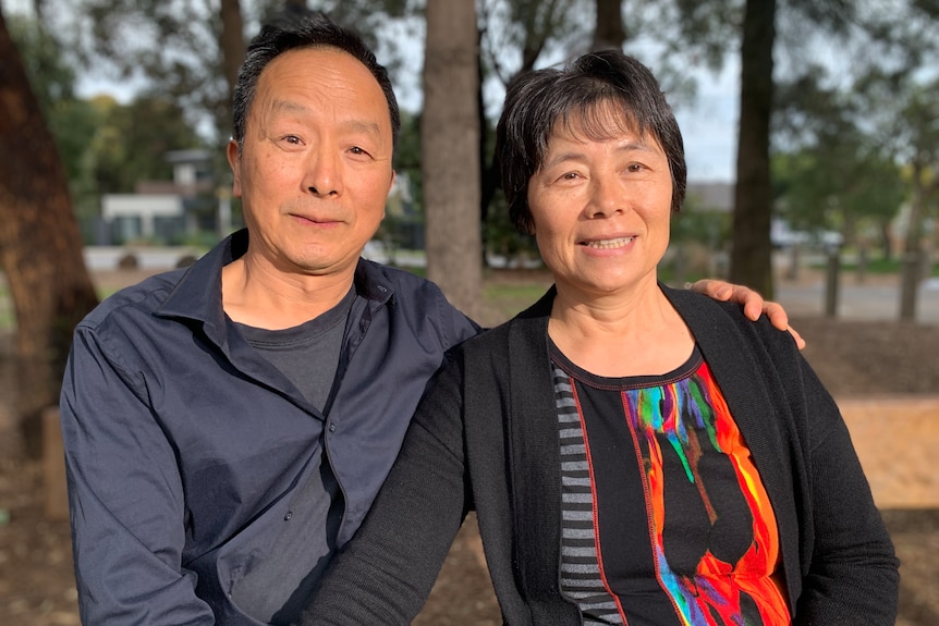 A Chinese couple smiling with the man's hand on the woman's shoulder and trees in the background.