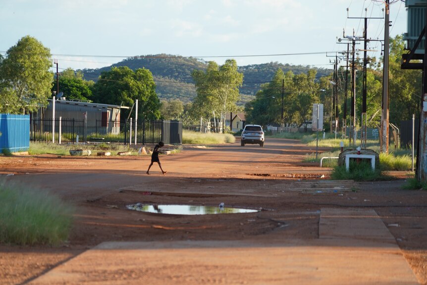 A child walking across a red dirt road that runs between houses, in a remote community.