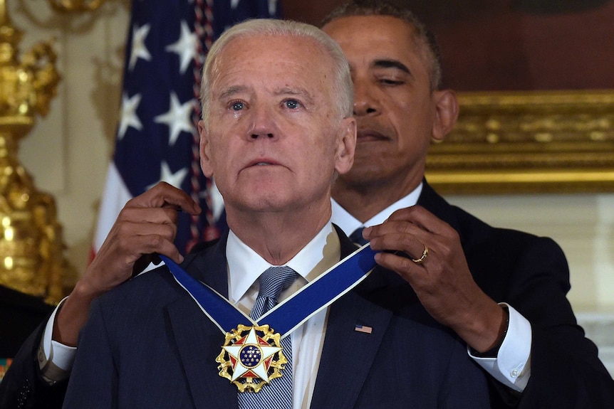 Joe Biden looks ahead with tears in his eyes as Barack Obama drapes a medal around his neck.
