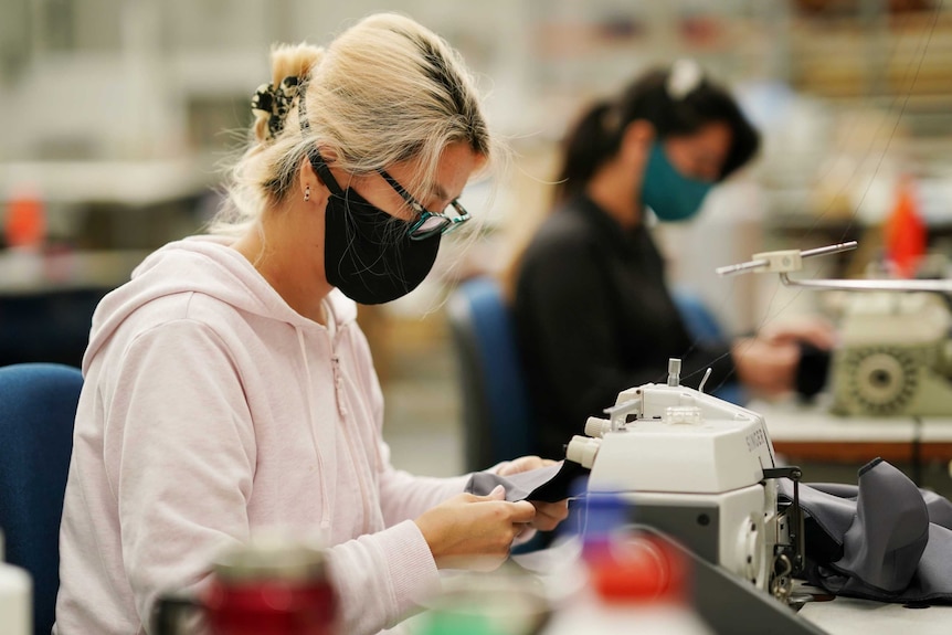 A woman, wearing a face mask, sits as a sewing machine with black fabric.