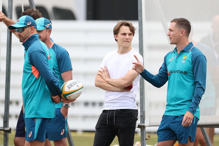 A former AFL player stands gesturing with his hand talking to a Formula One driver near Australian cricketers at a net session.