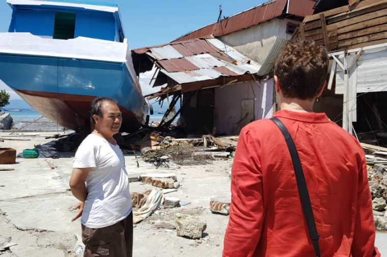 Journalist interview man in front of large fishing boat which was swept into a building on the beach.