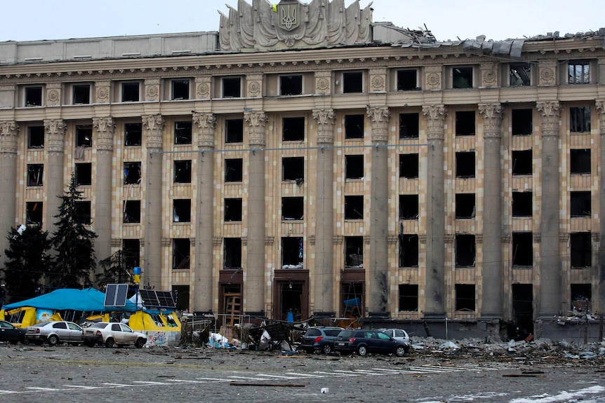 A stone building with an ornate feature on the roof is seen with significant damage, including windows and doors blown out