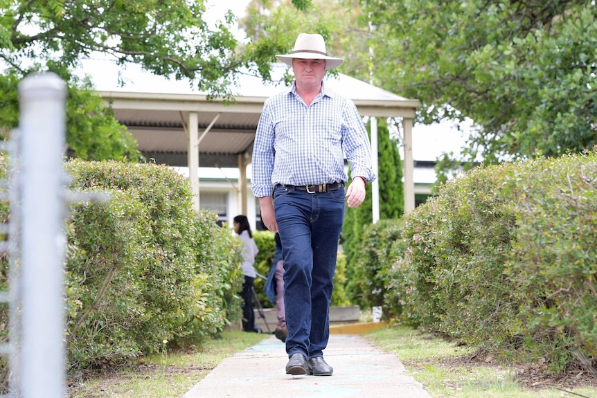 Barnaby Joyce walks down a path with a stern expression on his face. He is wearing blue jeans, a checked shirt and an akubra
