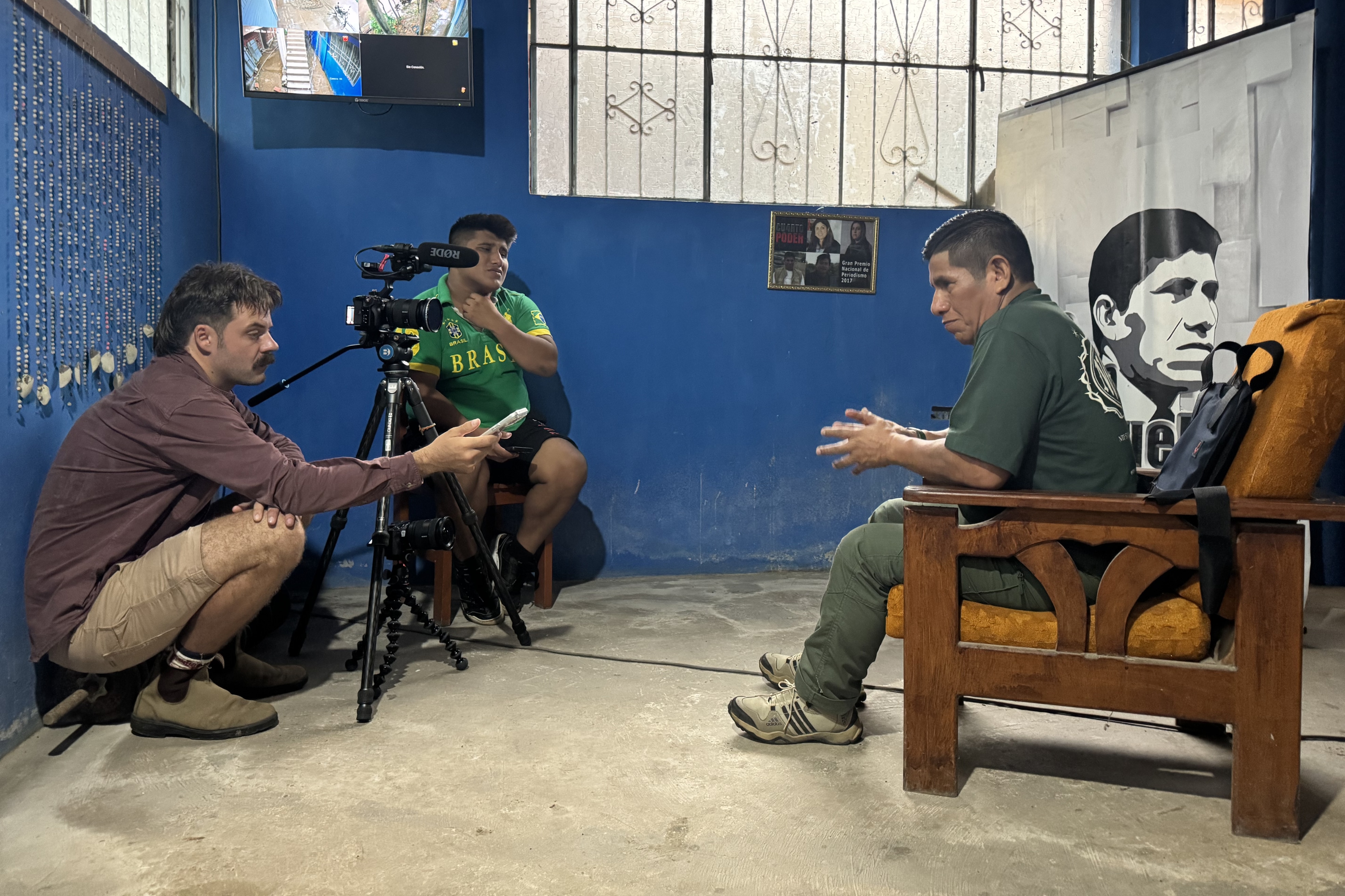 Jayden hunches on the ground, recording Manuel sitting in a chair, speaking.