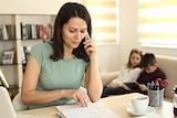 A woman peruses a spreadsheet while holding a phone to her ear. A pair of kids sit on a lounge behind her.