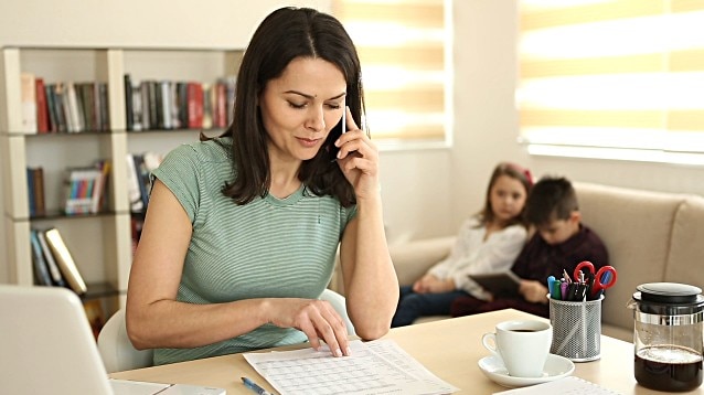 A woman peruses a spreadsheet while holding a phone to her ear. A pair of kids sit on a lounge behind her.