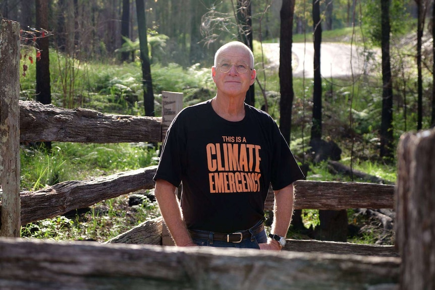 Man standing behind wooden fence of gravesite on his bush property looking up, delicate sunlight behind, wearing a climate shirt