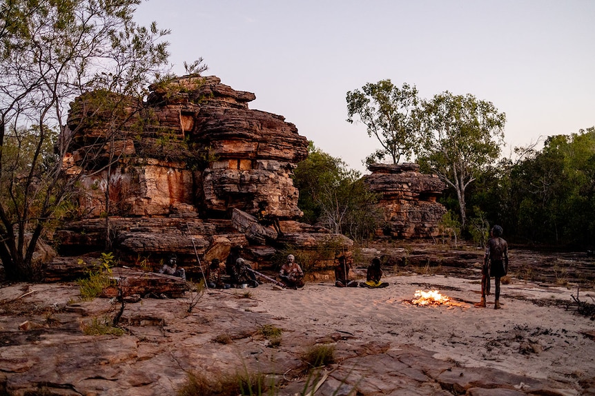 Six men sit and stand on the sand around a campfire in front of a rock shelf, holding didgeridoos and spears.