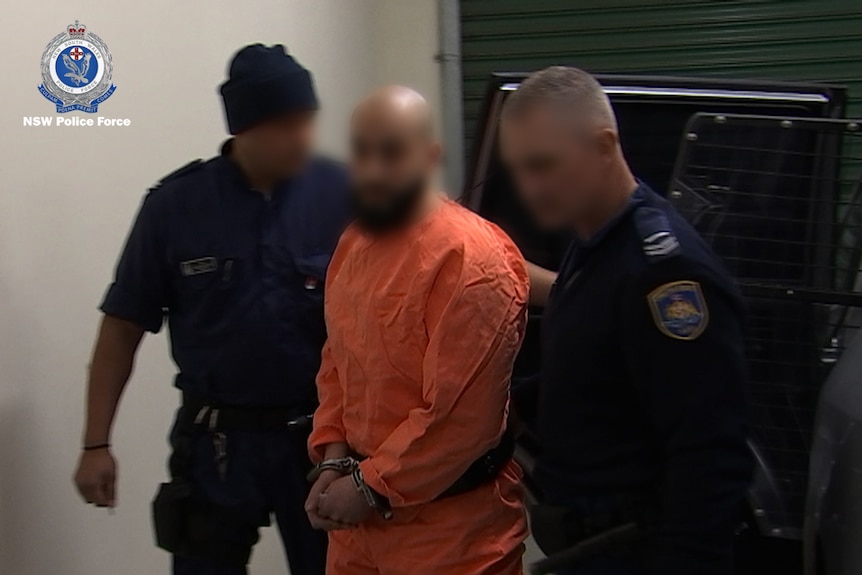 A man in an orange prison jumpsuit is flanked by two police officers