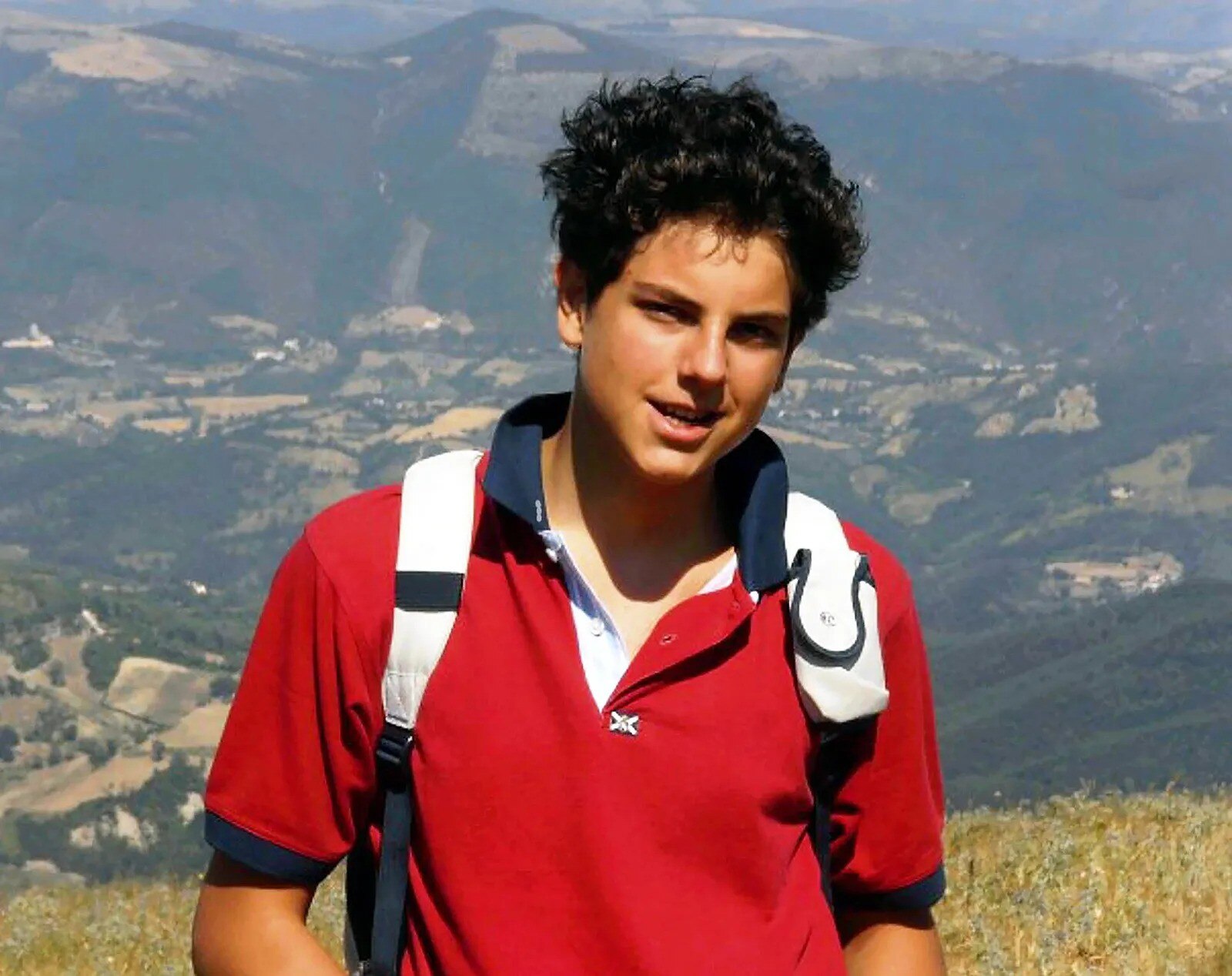 A teenager boy with olive skin and thick black hair wears a red polo shirt and white backpack and smiles.