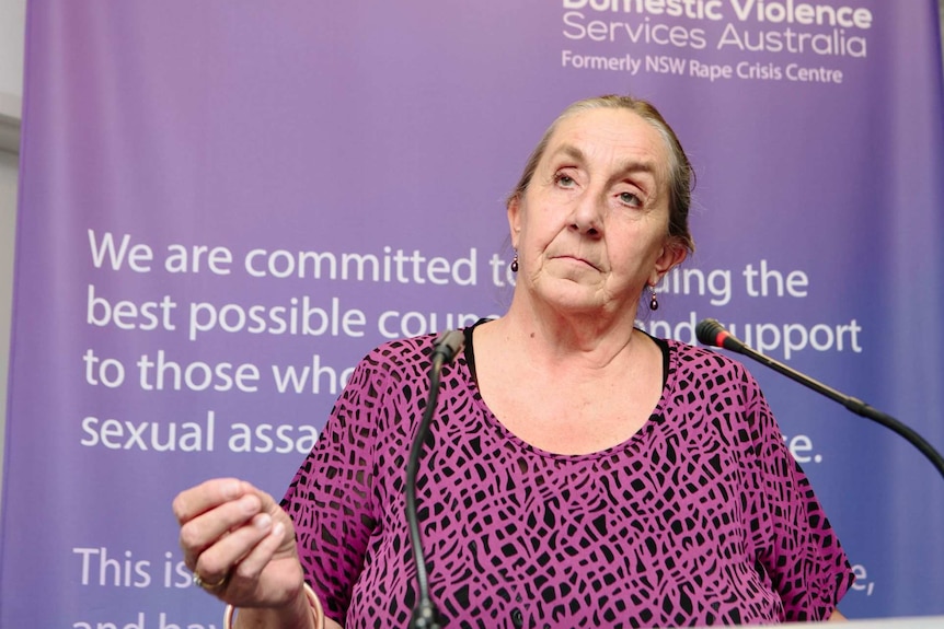 A woman with a serious expression and a black and purple top stands at a lectern.