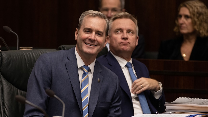 A man in the suit on the left looks happy with himself and a man in the right in a suit looks up up at the gallery 