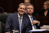 A man in the suit on the left looks happy with himself and a man in the right in a suit looks up up at the gallery 