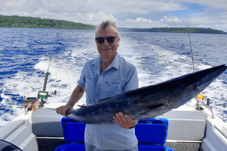 A man holds up a fish on the back of a boat in the Pacific with islands in the background