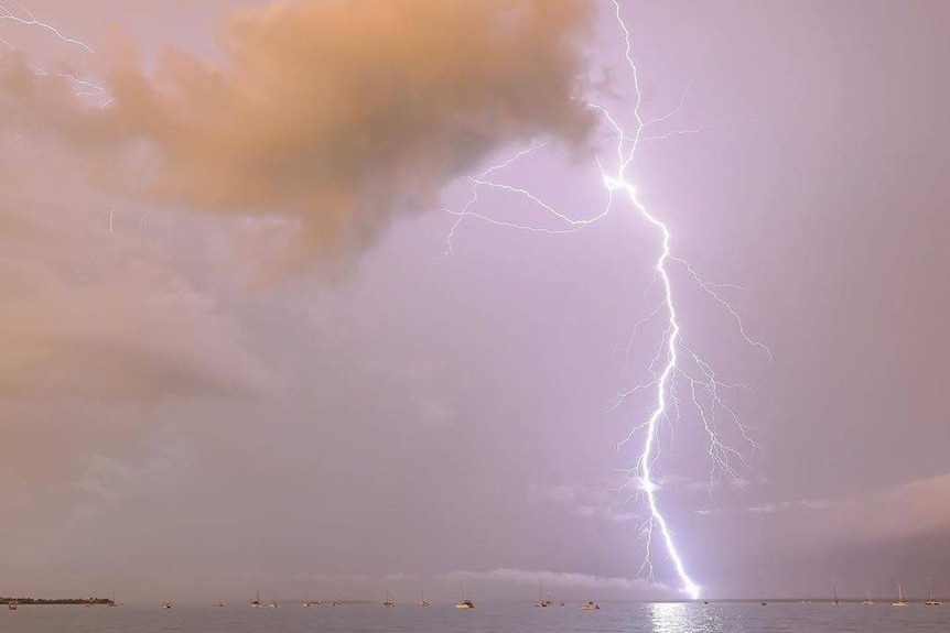 A lightning fork cracks over the ocean. There are boats out to sea, and red rocks in the foreground.
