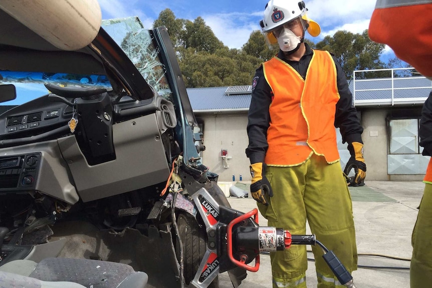 A firefighter wearing a mask and helmet watches on as a power tool forces a car apart.