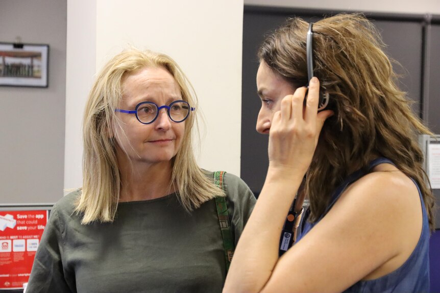 A blonde woman wearing blue glasses looks at a woman wearing a set of headphones.
