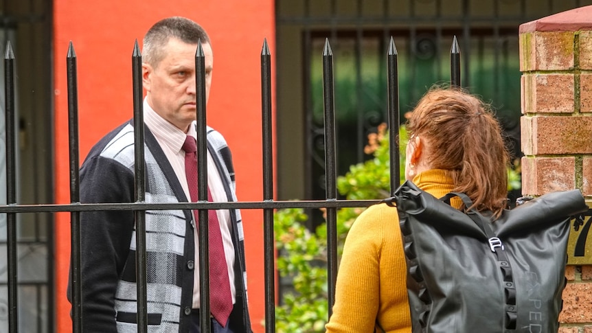 A man and woman stand on opposite sides of a closed gate, the woman's back the camera, the man glaring at her.