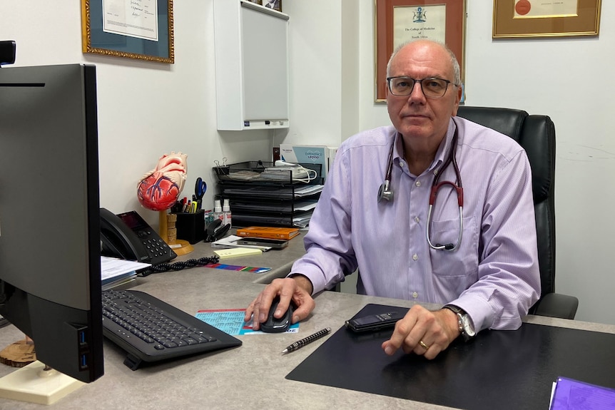 Bald unsmiling older man sits at desk with heart model, computer, lavender shirt, stethoscope around his neck, diplomas on wall.