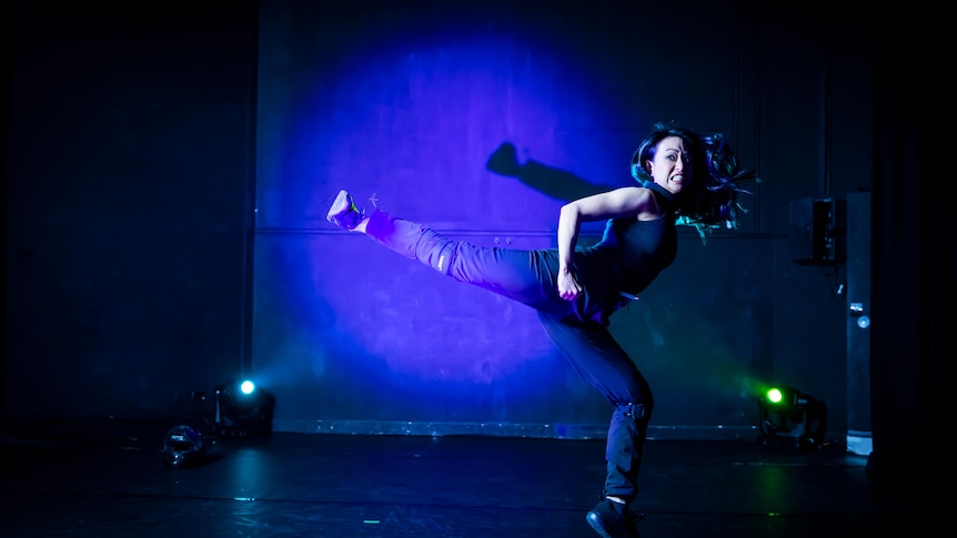 An Asian Australian woman does a flying kick on stage. She is lit by a blue light and wearing all black.