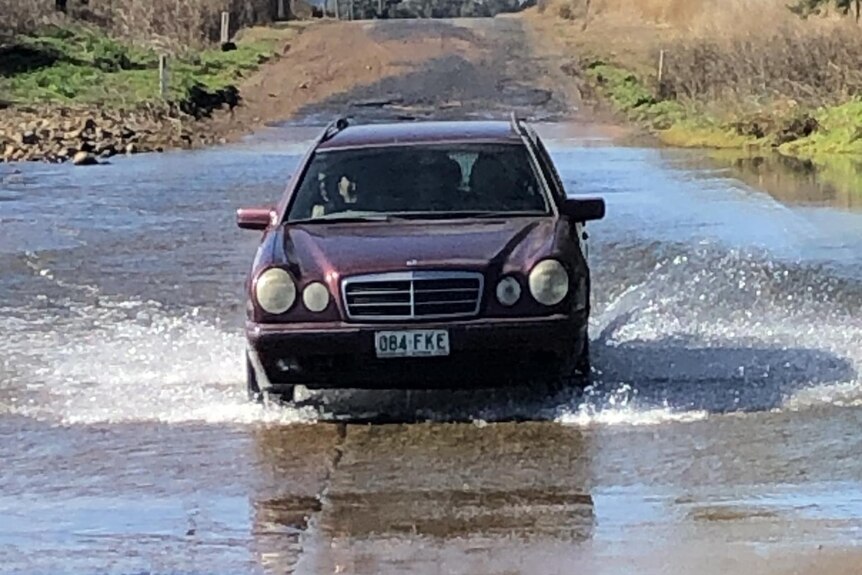 A maroon-coloured Mercedes Benz station wagon driving over a causeway that has water flowing over it