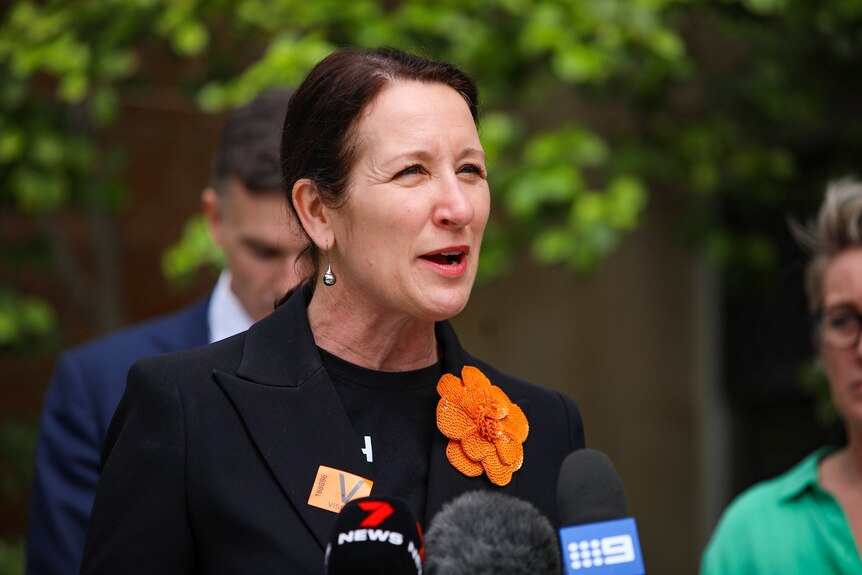 A woman with a black jacket and orange flower broche speaking at a press conference
