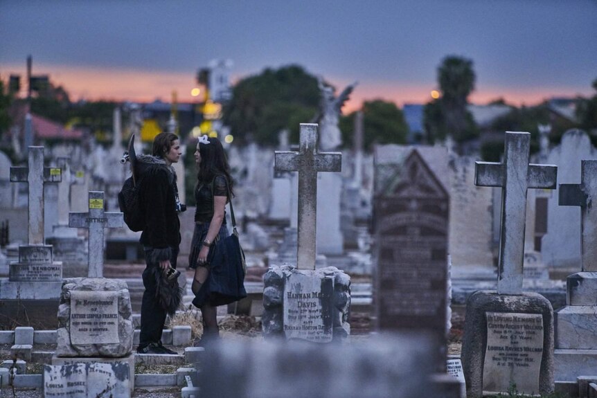A teenage couple stand in a cemetery as the sun sets.