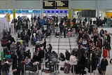 Dozens of people dressed in thick winter clothing stand around inside an airport terminal.