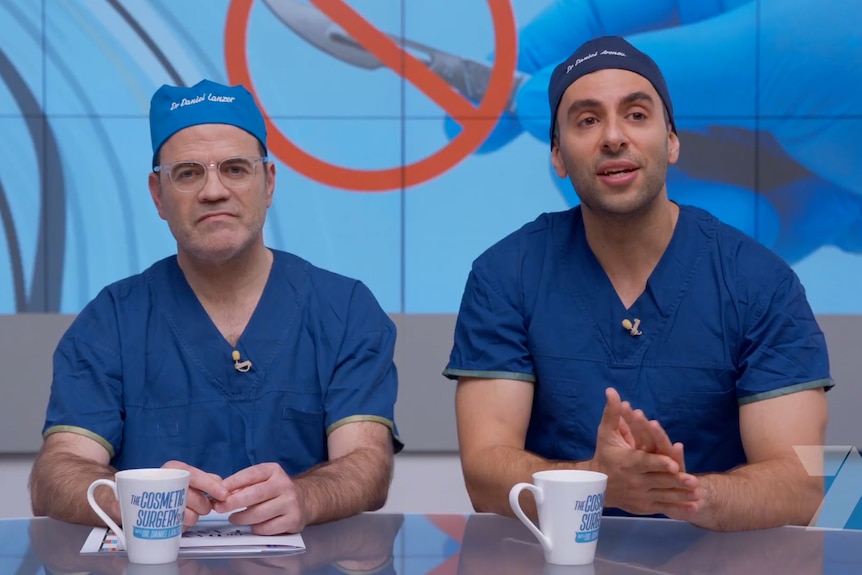 Two men in medical scrubs sit behind a desk on a TV set. 