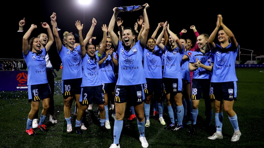 A group of W-League footballers cheers and raise their arms as a teammate lifts a trophy