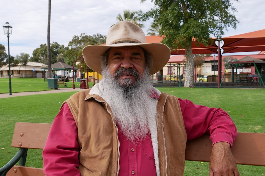 An Indigenous man with a long grey beard sitting on a park bench wearing a pink shirt, brown vest and a cowboy hat. 