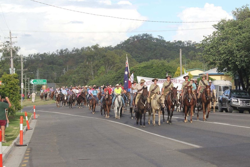 4  people in ANZAC uniform one holding an Australian flag lead a parade of horse riders through a Kilkivan street with onlookers