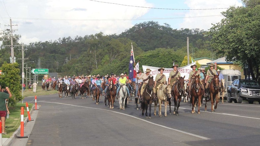 4  people in ANZAC uniform one holding an Australian flag lead a parade of horse riders through a Kilkivan street with onlookers