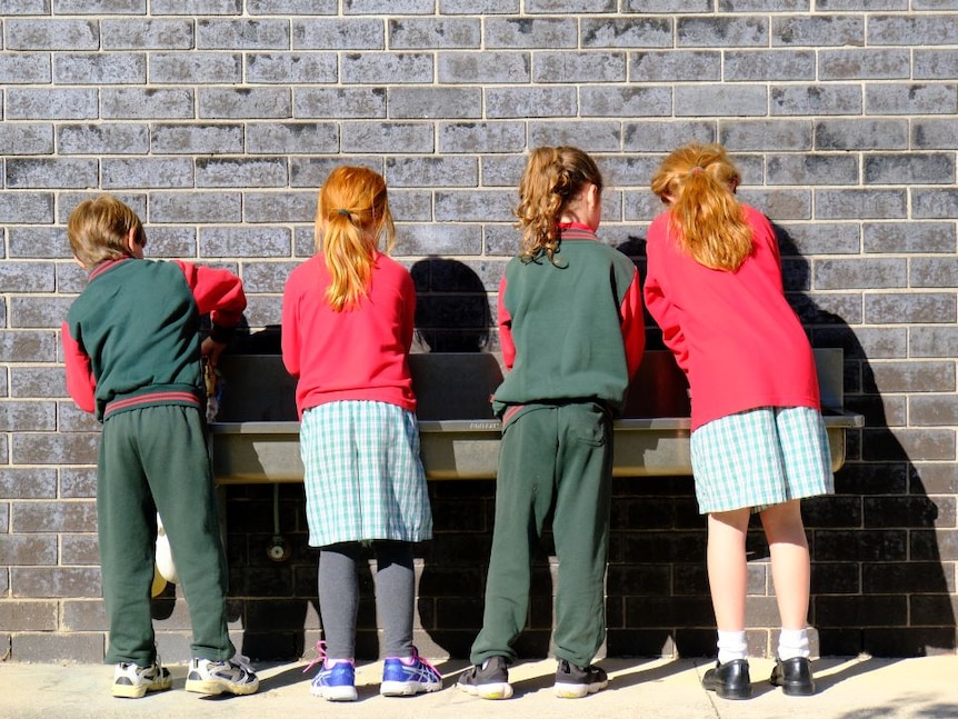 Four elementary school students stand with their backs to the camera and wash their hands in a long metal basin on a brick wall.