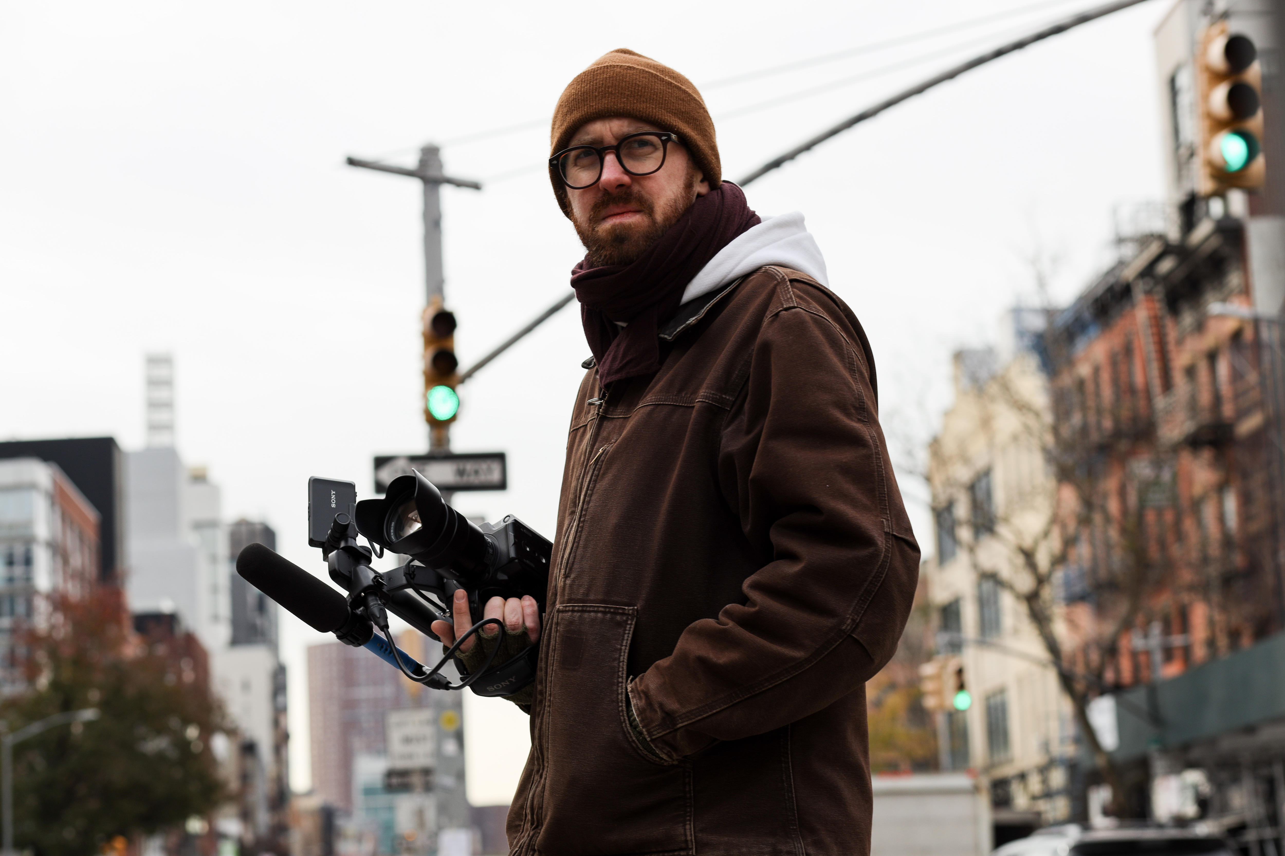 John Wilson stands looking concerned and rugged up on a NYC street, camera in hand.