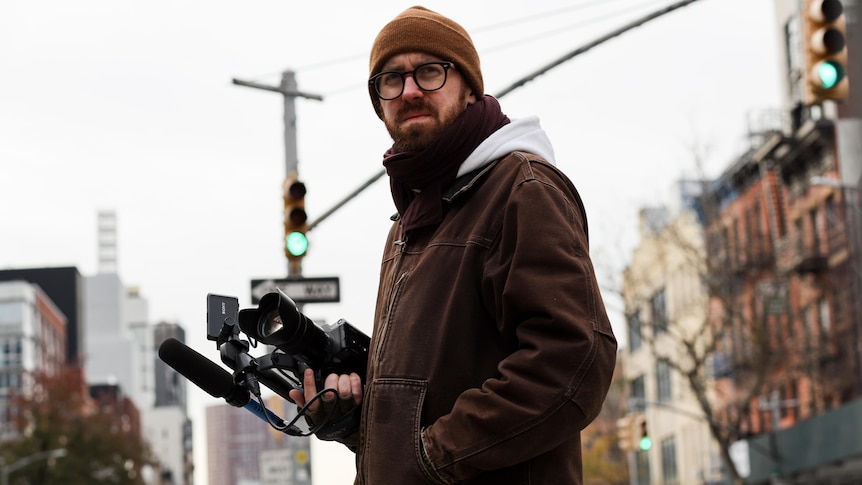 John Wilson stands looking concerned and rugged up on a NYC street, camera in hand.
